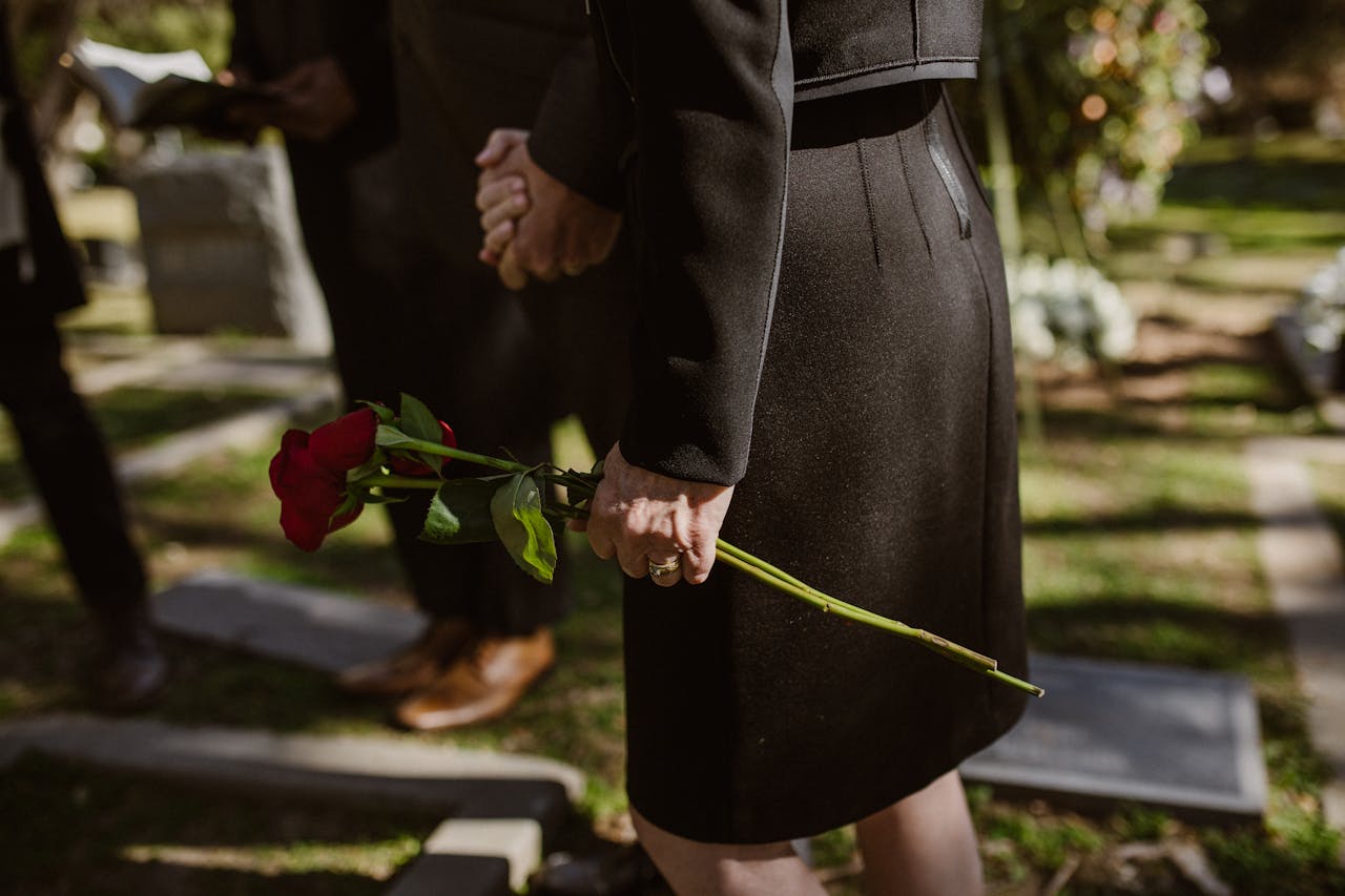 heros-img Close-up of a person in black attire holding a red rose at a cemetery.